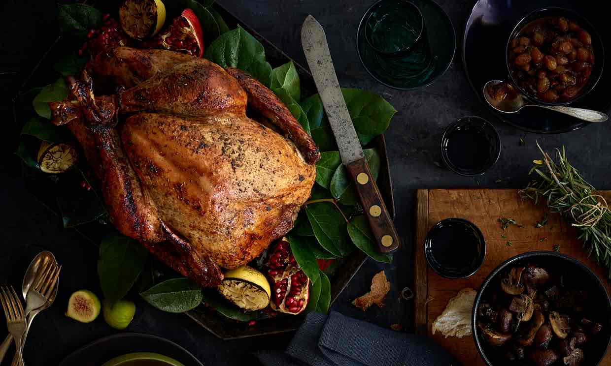 A roasted turkey on a platter with herbs, pomegranate, and lemon, beside side dishes.