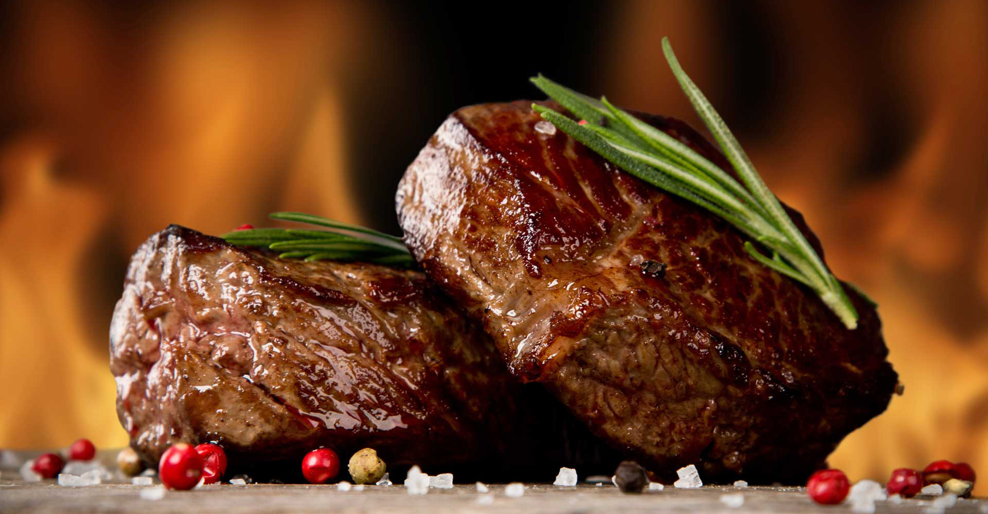 Two cooked steaks garnished with rosemary, surrounded by colorful peppercorns and salt.